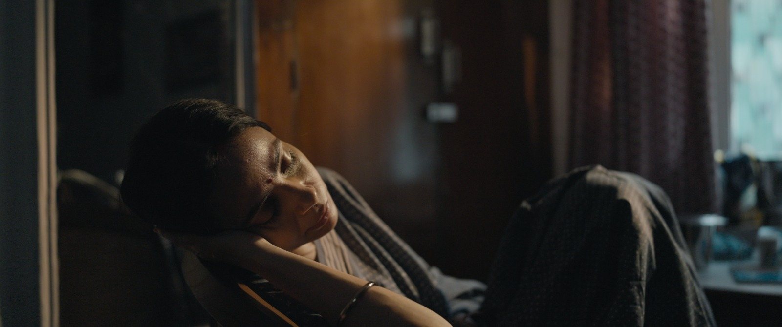 Kavita asleep in a chair in a darkened room, still dressed, beside the bed she has been tending.
