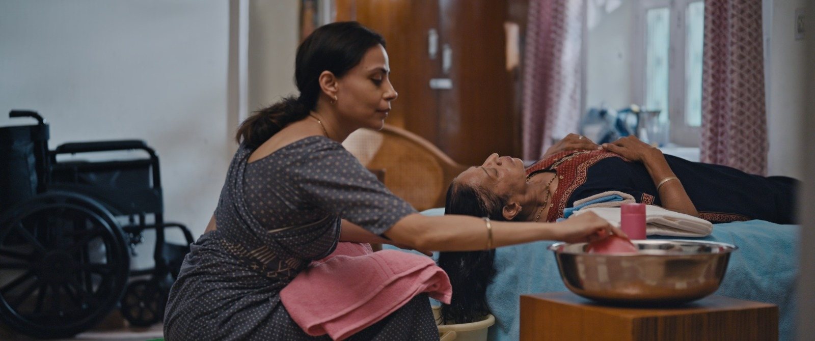 Kavita leans over a bed to tend to her mother-in-law, a wheelchair standing beside them.