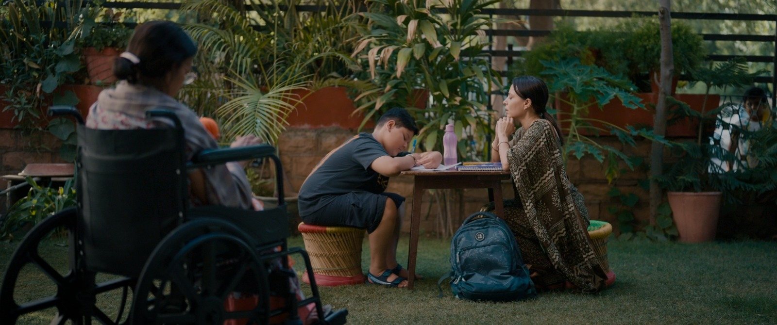 In a garden, an elderly woman in a wheelchair, a boy at a table and a woman seated on the ground — three generations together.