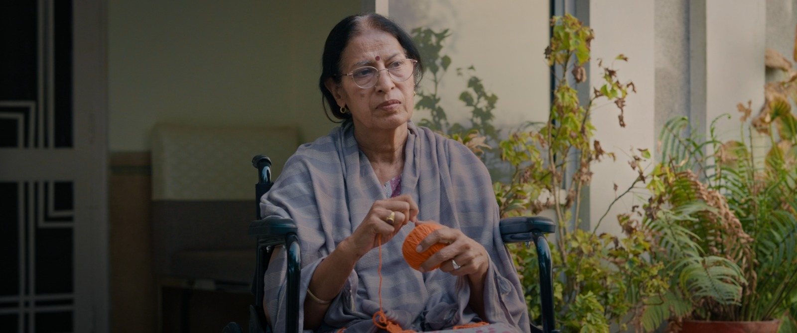 An older woman in a wheelchair on a verandah, peeling fruit, surrounded by plants.