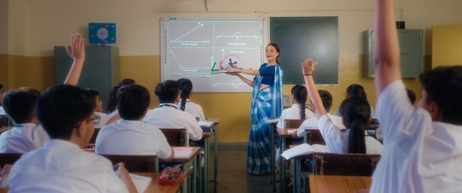 Kavita stands at the front of a classroom beside a projected lesson as her students raise their hands.